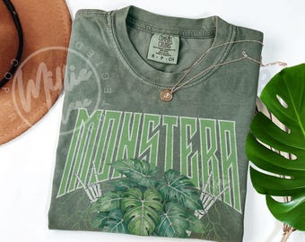 May include: Olive green t-shirt with the word "MONSTERA" in a green, outlined font. The design includes a monstera leaf graphic and skeleton hands. A gold necklace with a pendant is on the shirt.