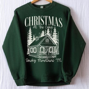 May include: A dark green sweatshirt with a white graphic of a cabin in the woods. The text "CHRISTMAS At the Cabin Smoky Mountains TN" is printed above and below the cabin illustration. The sweatshirt is hanging on a wooden hanger.