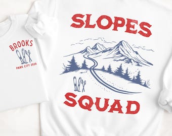 Puede incluir: Sudaderas blancas con gráficos de esquí. Una presenta "Brooks Park City 2026" con esquís. La otra muestra "Slopes Squad" con una escena de montaña y esquís. Ambas tienen diseños rojos y azules.