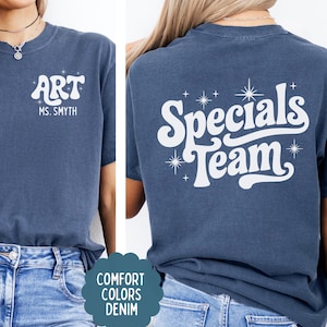 以下が含まれることがあります： デニムブルーのTシャツで、白いスクリーンプリントの文字が特徴です。前面には「ART」と「MS. SMYTH」の文字と小さな星のアクセントがあります。背面には、大きなスタイリッシュなフォントで「Specials Team」という文字が書かれ、いくつかの星のアイコンで囲まれています。