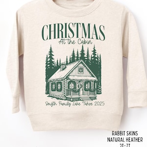 May include: A natural heather sweatshirt with a green graphic that reads "CHRISTMAS At the Cabin" above a cabin illustration. Below the cabin is "Smyth Family Lake Tahoe 2025". The sweatshirt is labeled "RABBIT SKINS NATURAL HEATHER 2T-7T".