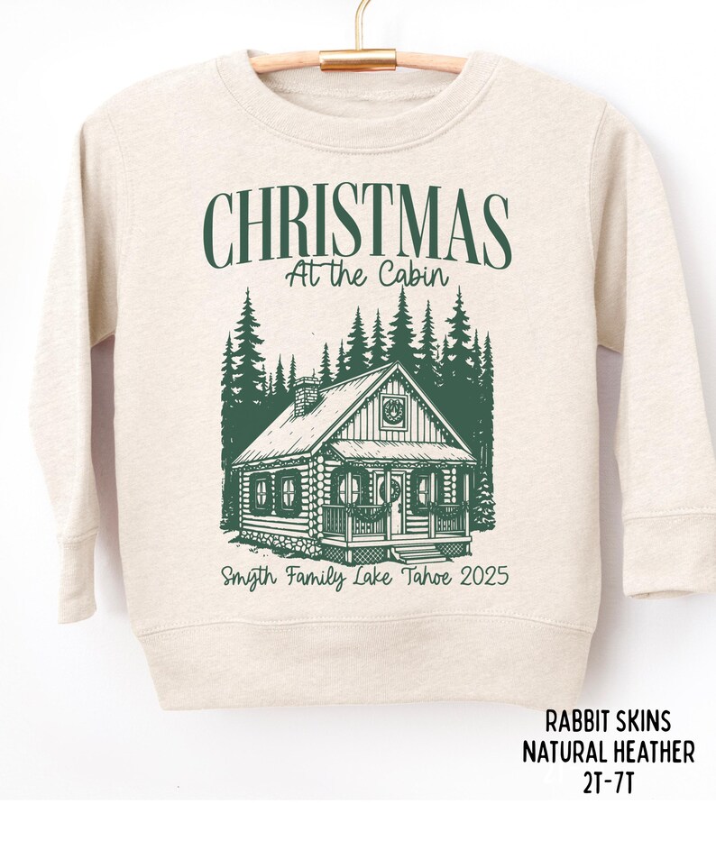 May include: A natural heather sweatshirt with a green graphic that reads "CHRISTMAS At the Cabin" above a cabin illustration. Below the cabin is "Smyth Family Lake Tahoe 2025". The sweatshirt is labeled "RABBIT SKINS NATURAL HEATHER 2T-7T".
