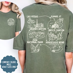 May include: Moss green t-shirt with a white graphic of a family crest and the text "SMYTH FAMILY AUGUST 2026" on the front. The back features a travel route map with landmarks like the Grand Canyon and Las Vegas. "Comfort Colors" and "Adult Sizes Only" are also displayed.