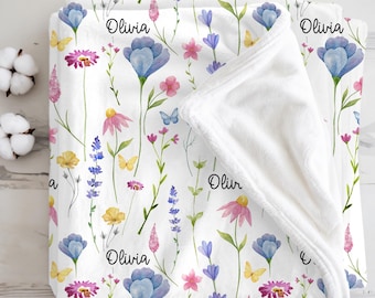 May include: White floral blanket featuring a watercolor design of blue, pink, yellow, and purple flowers, with butterflies. The name "Olivia" is printed in black script across the blanket. The blanket is folded over, revealing a soft, white interior.
