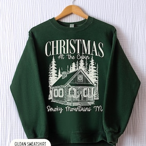 May include: Forest green sweatshirt with a white graphic of a cabin in the woods, with the text "CHRISTMAS At the Cabin Smoky Mountains TN". The sweatshirt is displayed on a wooden hanger.