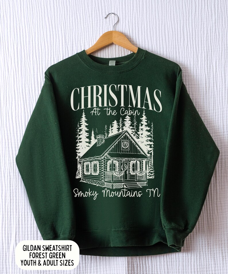 May include: Forest green sweatshirt with a white graphic of a cabin in the woods, with the text "CHRISTMAS At the Cabin Smoky Mountains TN". The sweatshirt is displayed on a wooden hanger.