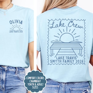 May include: A light blue t-shirt featuring a dark blue graphic. The front chest displays the name OLIVIA above a sun and the text LAKE TRAVIS 2026. The back features a large stamp-shaped design with a dock, sun, and text LAKE CREW SMYTH FAMILY 2026.