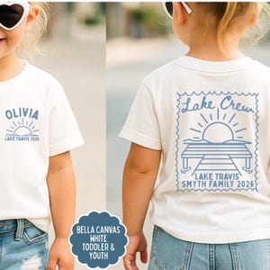 May include: A white short-sleeved t-shirt featuring blue graphics. The front displays the name OLIVIA above a sun icon and the text LAKE TRAVIS 2026. The back shows a stamp-shaped design with a dock, sun, and the text Lake Crew LAKE TRAVIS SMYTH FAMILY 2026.