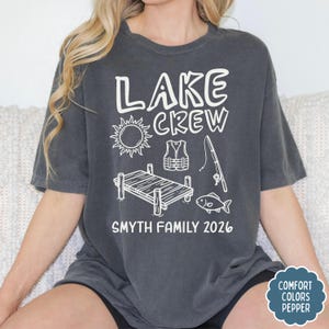 May include: Dark gray t-shirt with the words "LAKE CREW" and a sun graphic, life vest, fishing rod, dock, and fish. Below the graphics is "SMYTH FAMILY 2026". The shirt has a Comfort Colors Pepper tag.