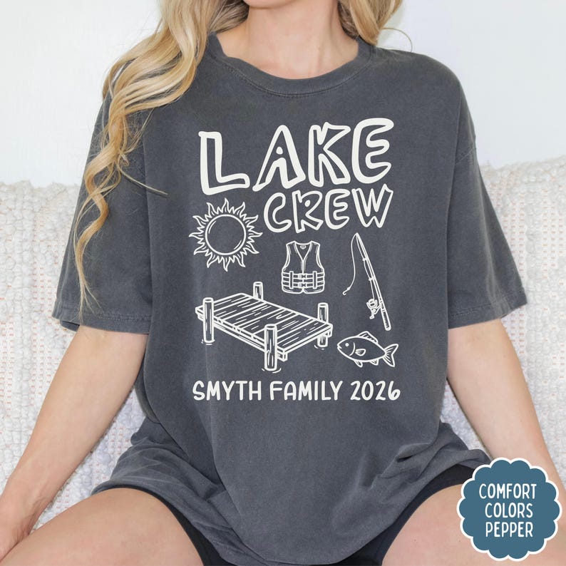May include: Dark gray t-shirt with the words "LAKE CREW" and a sun graphic, life vest, fishing rod, dock, and fish. Below the graphics is "SMYTH FAMILY 2026". The shirt has a Comfort Colors Pepper tag.