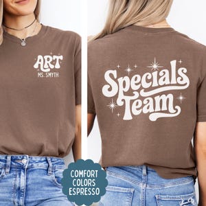 以下が含まれることがあります： 白い文字がプリントされた茶色の半袖Tシャツ。左胸には「ART」と「MS. SMYTH」の文字。背面には大きなフォントで「Specials Team」という文字と星のアクセントがデザインされています。Tシャツには「Comfort Colors Espresso」のラベルが付いています。