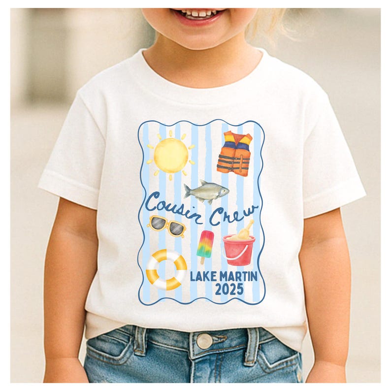 May include: White t-shirt featuring a blue and white striped design with beach-themed illustrations, including a sun, life vest, fish, sunglasses, popsicle, bucket, and a life preserver. The text "Cousin Crew Lake Martin 2025" is also displayed.