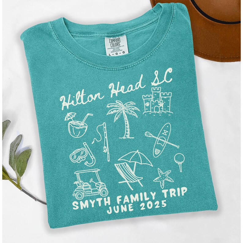 May include: Teal blue t-shirt with a white graphic design of beach vacation icons and the text "Hilton Head SC Smyth Family Trip June 2025".