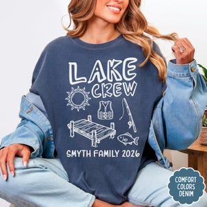 May include: Navy blue sweatshirt with white text and graphics. The text reads "LAKE CREW" above a sun, life vest, fishing rod, dock, and fish. Below is "SMYTH FAMILY 2026". The sweatshirt is part of the Comfort Colors Denim collection.