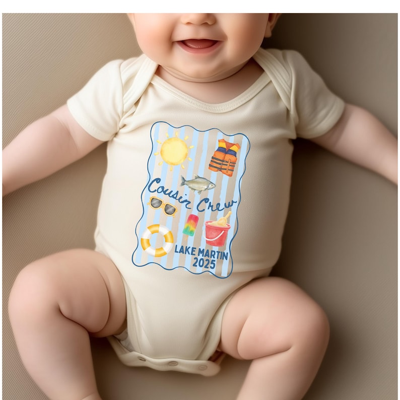 May include: A cream-colored baby onesie with a colorful graphic that reads "Cousin Crew Lake Martin 2025." The graphic includes a sun, sunglasses, a fish, a life vest, a popsicle, a bucket, and a life preserver.