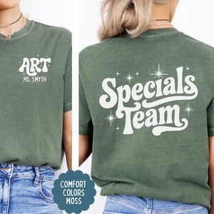 以下が含まれることがあります： モスグリーンのTシャツで、白い文字がプリントされています。前面には「ART」と「MS. SMYTH」の文字があります。背面には大きなスタイリッシュなフォントで「Specials Team」と書かれており、小さな星のグラフィックが周囲に配置されています。青いデニムジーンズと組み合わされています。