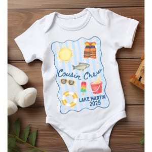 May include: White baby onesie with a blue and yellow graphic. The graphic includes a sun, life vest, fish, sunglasses, popsicle, bucket of ice cream, and a life preserver. Text reads "Cousin Crew Lake Martin 2025."