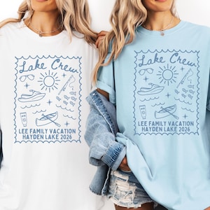 May include: White and light blue t-shirts with a blue graphic design. The design includes the text "Lake Crew" and illustrations of sunglasses, a sun, a jet ski, fish, sunscreen, and a canoe. The text "LEE FAMILY VACATION HAYDEN LAKE 2026" is also included.