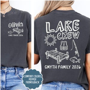 May include: Dark gray t-shirt with white graphic designs. The front features the name "Olivia", a fish, and a dock, with "LAKE TAHOE 2026" below. The back displays "LAKE CREW", a sun, life vest, fishing rod, dock, fish, and "SMYTH FAMILY 2026".