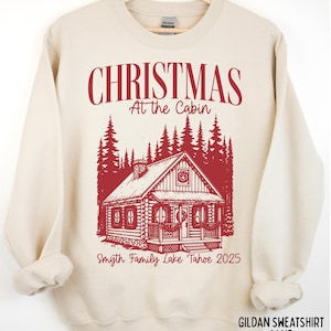 May include: Sand-colored sweatshirt with a red graphic of a cabin in a forest, with the text "CHRISTMAS At the Cabin" and "Smyth Family Lake Tahoe 2025". The sweatshirt is made of Gildan material.