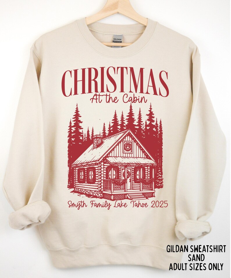 May include: Sand-colored sweatshirt with a red graphic of a cabin in a forest, with the text "CHRISTMAS At the Cabin" and "Smyth Family Lake Tahoe 2025". The sweatshirt is made of Gildan material.