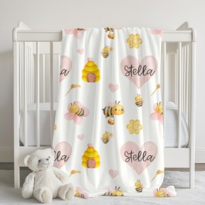 May include: White baby blanket with a repeating pattern of bees, honeycombs, pink hearts, and beehives. The name "Stella" is written in script on two of the hearts. A teddy bear sits in front of a white crib.