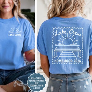 May include: A heather Columbia blue t-shirt featuring a white graphic of a sun over Lake Tahoe on the front chest. The back displays a large white stamp-shaped graphic with a dock, sun, and the text Lake Crew HOMEWOOD 2026 in white ink.