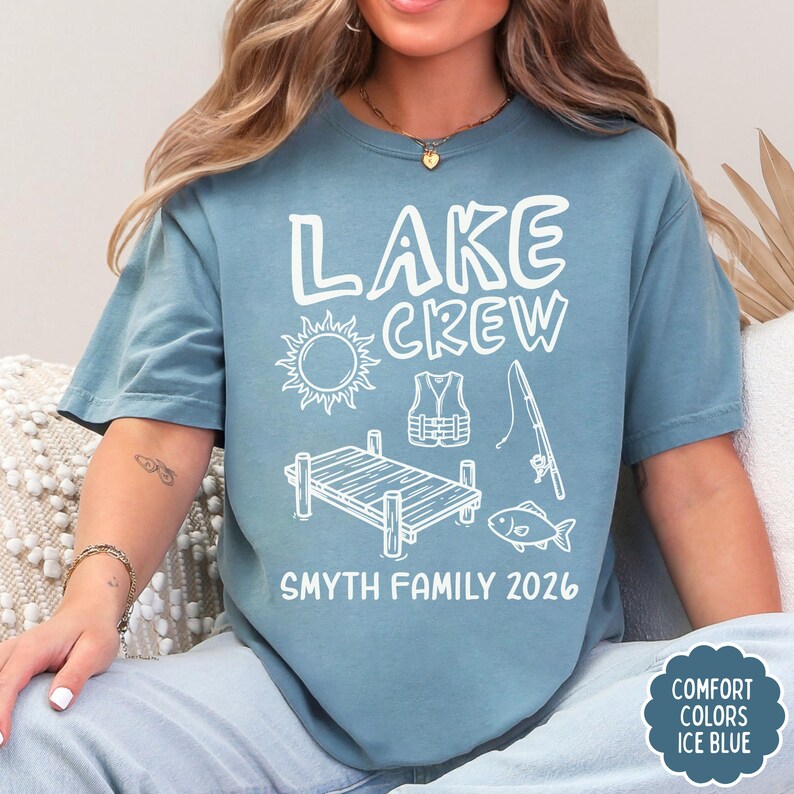 May include: A light blue t-shirt with the words "LAKE CREW" and a graphic of a sun, dock, life vest, fishing pole, and fish. The text "SMYTH FAMILY 2026" is below the graphic. The shirt is labeled "COMFORT COLORS ICE BLUE".