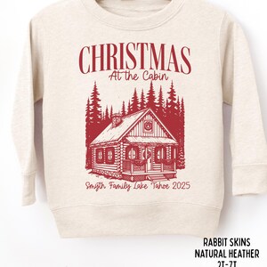 May include: A natural heather sweatshirt with a red graphic. The graphic features the text "CHRISTMAS At the Cabin" above a cabin illustration and the text "Smyth Family Lake Tahoe 2025".