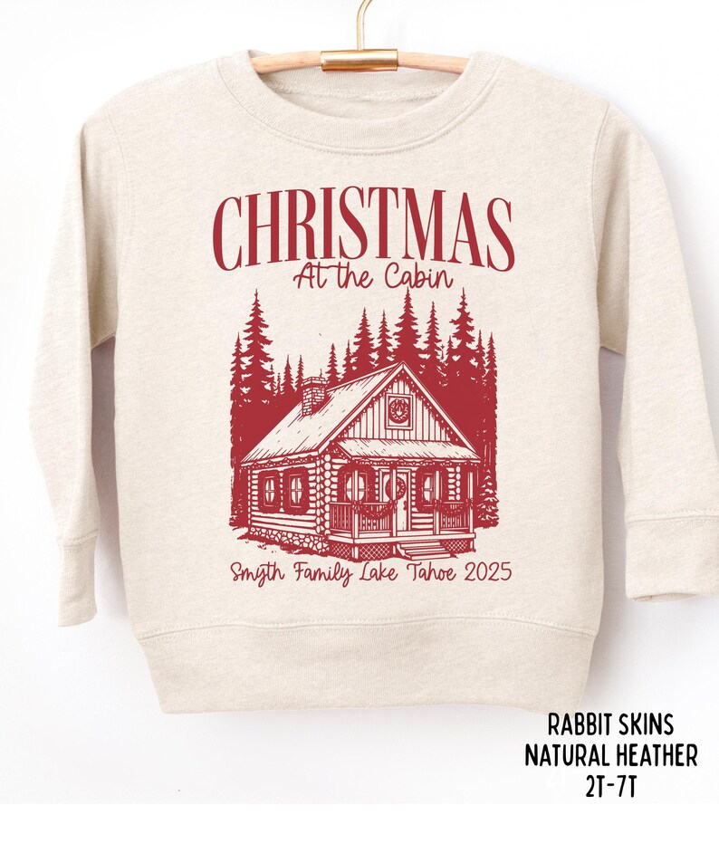 May include: A natural heather sweatshirt with a red graphic. The graphic features the text "CHRISTMAS At the Cabin" above a cabin illustration and the text "Smyth Family Lake Tahoe 2025".
