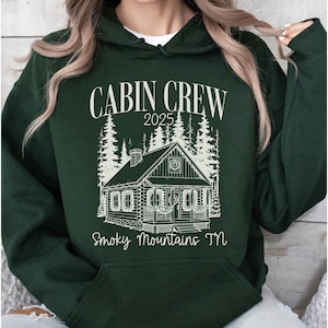 May include: Dark green hoodie with a white graphic that reads "CABIN CREW 2025" above a cabin illustration and "Smoky Mountains TN" below. The cabin is surrounded by trees. The hoodie has a front pocket and a hood.