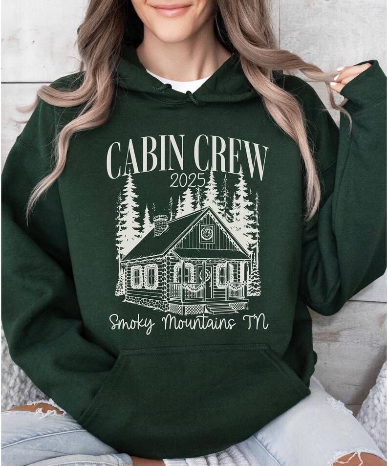 May include: Dark green hoodie with a white graphic that reads "CABIN CREW 2025" above a cabin illustration and "Smoky Mountains TN" below. The cabin is surrounded by trees. The hoodie has a front pocket and a hood.