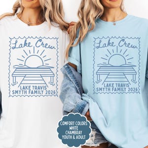 May include: A white t-shirt and a light blue t-shirt, each featuring a blue graphic of a sun rising over a dock inside a postage stamp border. The text reads Lake Crew, Lake Travis, and Smyth Family 2026. A label notes Comfort Colors, White, Chambray.