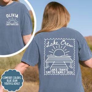 May include: A blue jean coloured t-shirt featuring white screen-printed graphics. The back displays a stamp-shaped design with a sun, a dock, and the text Lake Crew, Lake Travis, Smyth Family 2026. The front chest shows Olivia and Lake Travis 2026.