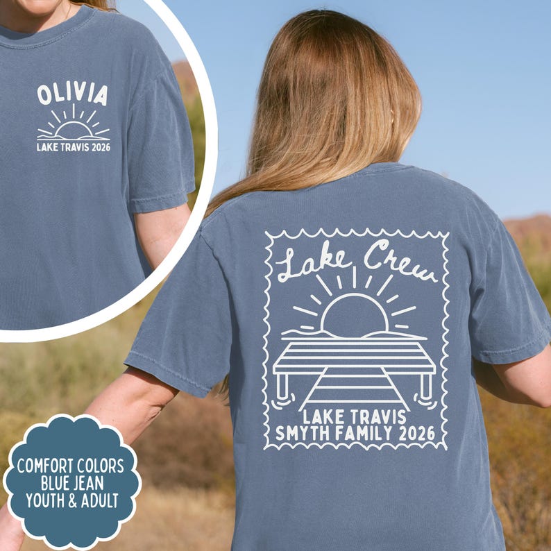 May include: A blue jean coloured t-shirt featuring white screen-printed graphics. The back displays a stamp-shaped design with a sun, a dock, and the text Lake Crew, Lake Travis, Smyth Family 2026. The front chest shows Olivia and Lake Travis 2026.