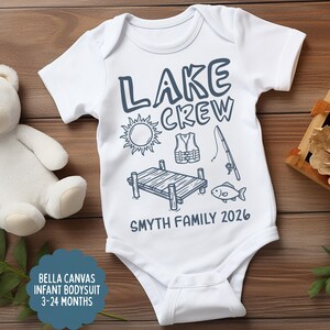 May include: White infant bodysuit with the text "LAKE CREW" and lake-themed illustrations in navy blue. Includes a sun, life vest, fishing rod, dock, and fish. The text "SMYTH FAMILY 2026" is also printed on the bodysuit.
