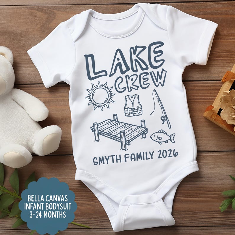 May include: White infant bodysuit with the text "LAKE CREW" and lake-themed illustrations in navy blue. Includes a sun, life vest, fishing rod, dock, and fish. The text "SMYTH FAMILY 2026" is also printed on the bodysuit.