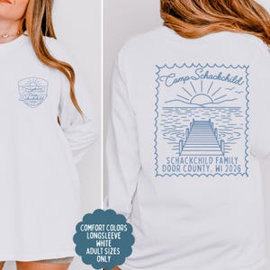 May include: A white long-sleeve shirt featuring a blue graphic design. The front displays a small shield logo with a canoe and sun. The back features a large stamp-shaped graphic of a lake, dock, and sun with the text Camp Schackchild, Schackchild Family, Door County, WI 2026.