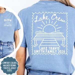 May include: A denim blue t-shirt featuring a white graphic on the back of a dock, sun, and the text Lake Crew, Lake Travis Smyth Family 2026. The front chest area displays the text Olivia, Lake Travis 2026 with a small sun graphic.