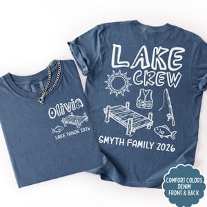 May include: Two denim blue t-shirts with white graphic designs. One shirt says "Olivia" and "Lake Tahoe 2026". The other says "Lake Crew" with a sun, pier, life vest, fishing rod, and fish. "Smyth Family 2026" is also printed.
