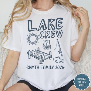 May include: White t-shirt with a navy blue graphic that says "LAKE CREW" with illustrations of a sun, life vest, fishing pole, dock, and fish. The text "SMYTH FAMILY 2026" is below the illustrations.