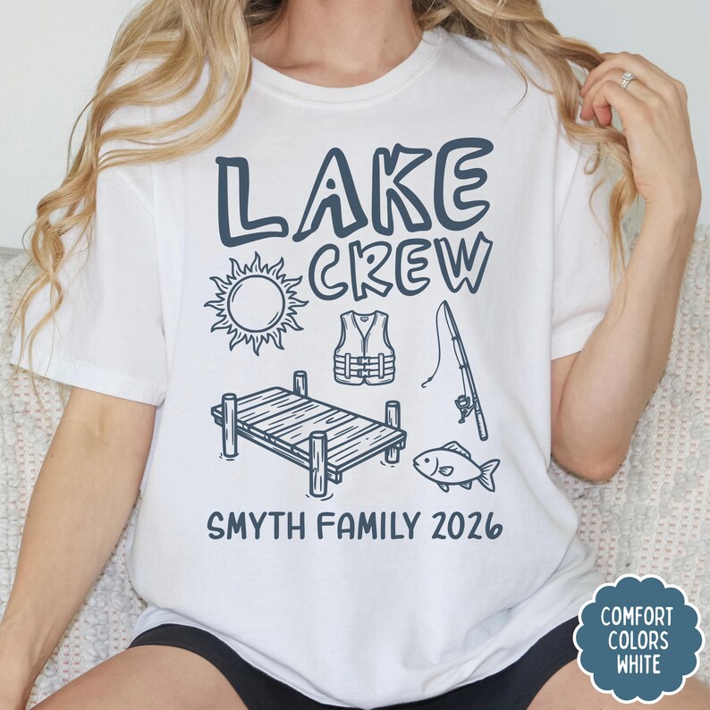 May include: White t-shirt with a navy blue graphic that says "LAKE CREW" with illustrations of a sun, life vest, fishing pole, dock, and fish. The text "SMYTH FAMILY 2026" is below the illustrations.