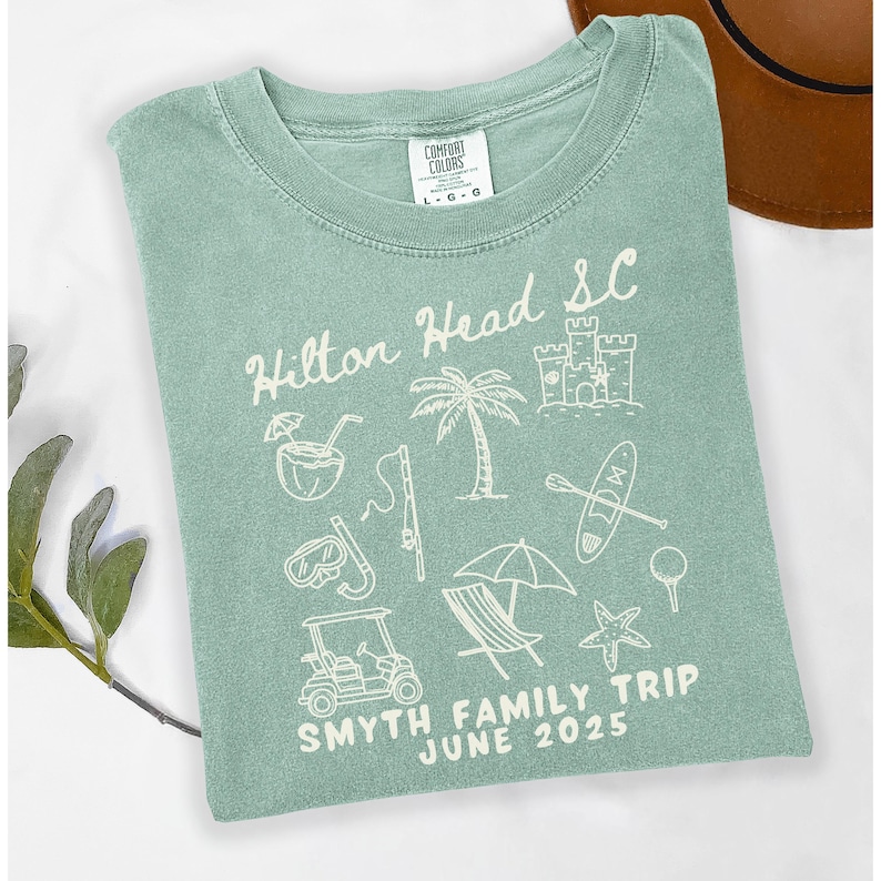May include: A light green t-shirt with a white graphic design featuring the text "Hilton Head SC" and various beach-themed icons, including a palm tree, a castle, a surfboard, a golf ball, a golf cart, and a starfish. The text "SMYTH FAMILY TRIP JUNE 2025" is also included in the design.