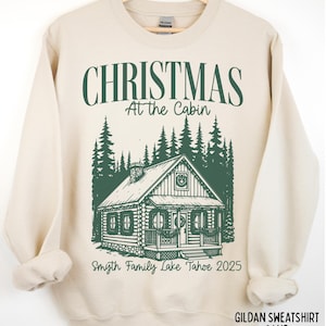 May include: Sand-colored sweatshirt with a green graphic. The graphic features the text "CHRISTMAS At the Cabin" above a cabin illustration and the text "Smyth Family Lake Tahoe 2025". The sweatshirt is hanging on a wooden hanger.