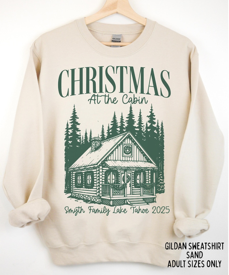 May include: Sand-colored sweatshirt with a green graphic. The graphic features the text "CHRISTMAS At the Cabin" above a cabin illustration and the text "Smyth Family Lake Tahoe 2025". The sweatshirt is hanging on a wooden hanger.