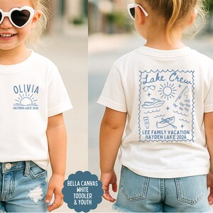 May include: White toddler t-shirt with a blue graphic on the front that says "OLIVIA" above a sun and the text "HAYDEN LAKE 2026". The back of the shirt has a blue "Lake Crew" design with vacation-themed illustrations and text.