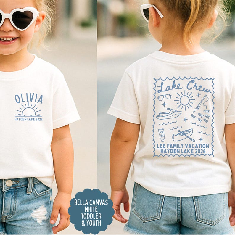 May include: White toddler t-shirt with a blue graphic on the front that says "OLIVIA" above a sun and the text "HAYDEN LAKE 2026". The back of the shirt has a blue "Lake Crew" design with vacation-themed illustrations and text.