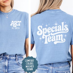 以下が含まれることがあります： 白のスクリーン印刷が施されたライトブルーのTシャツ。前面には「ART」と「MS. SMYTH」の文字と小さな星のアクセントがあります。背面には、大きなスタイリッシュなフォントで「Specials Team」という文字が描かれ、周囲に様々な星のアイコンが配置されています。