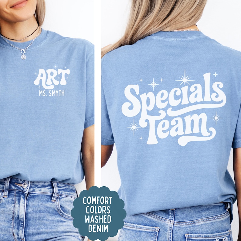 以下が含まれることがあります： 白のスクリーン印刷が施されたライトブルーのTシャツ。前面には「ART」と「MS. SMYTH」の文字と小さな星のアクセントがあります。背面には、大きなスタイリッシュなフォントで「Specials Team」という文字が描かれ、周囲に様々な星のアイコンが配置されています。