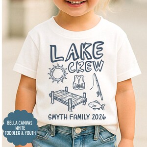 May include: White toddler and youth t-shirt with a navy blue graphic that says "LAKE CREW" with illustrations of a sun, life vest, fishing pole, dock, and fish. The text "SMYTH FAMILY 2026" is below the graphic.